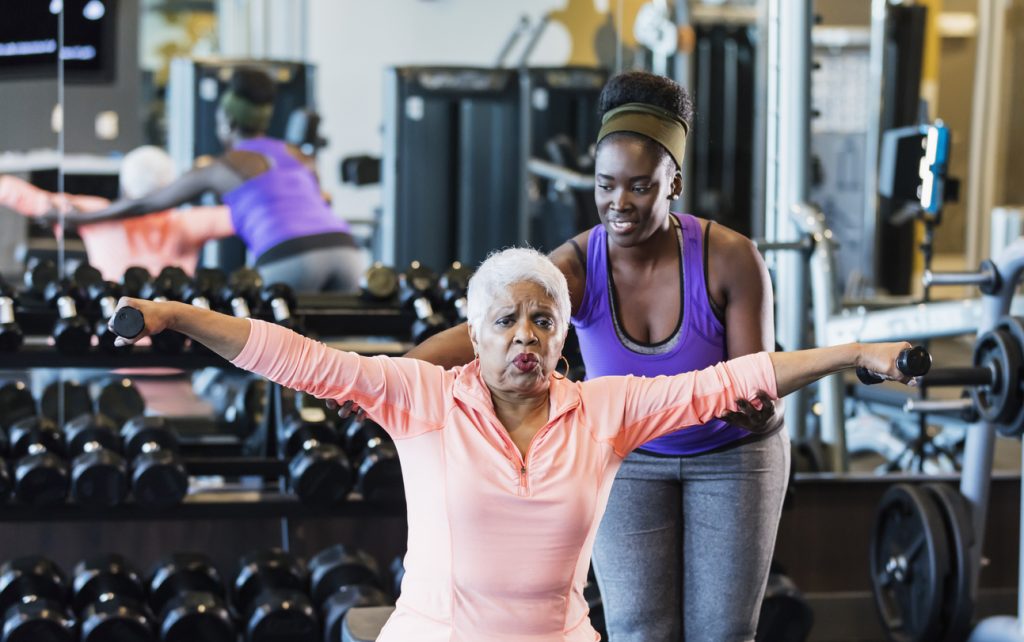 A young African-American woman in her 20s working as a personal trainer or fitness instructor in a gym. She is helping a senior African-American woman in her 60s who grimacing as she lifts hand weights.