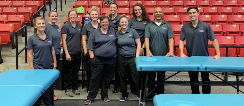 A group of students and teachers stand behind a few massage tables in a sports arena. 